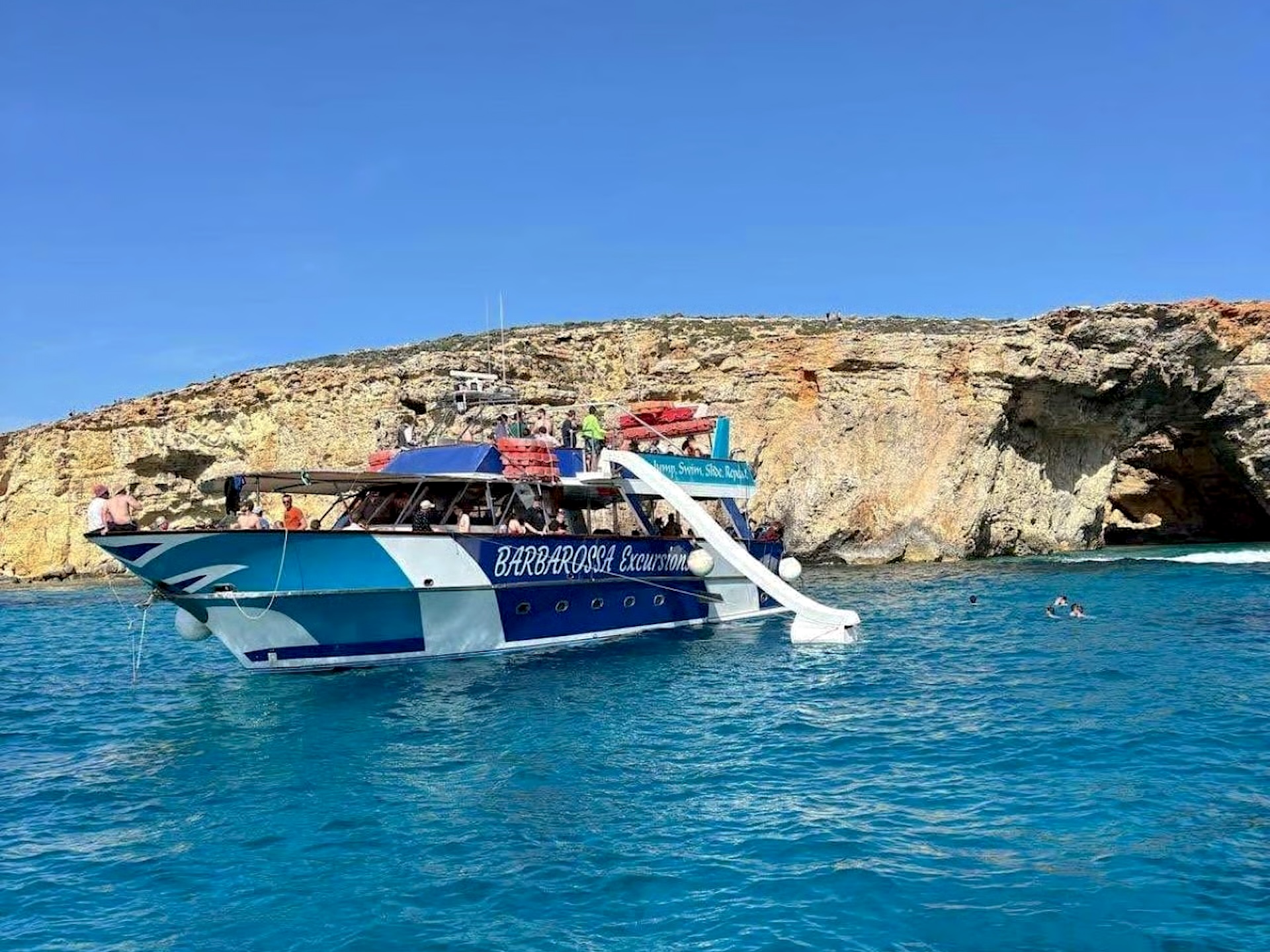 Escursione alla Blue Lagoon Barca blu e bianca con scivolo sulla riva di Comino
