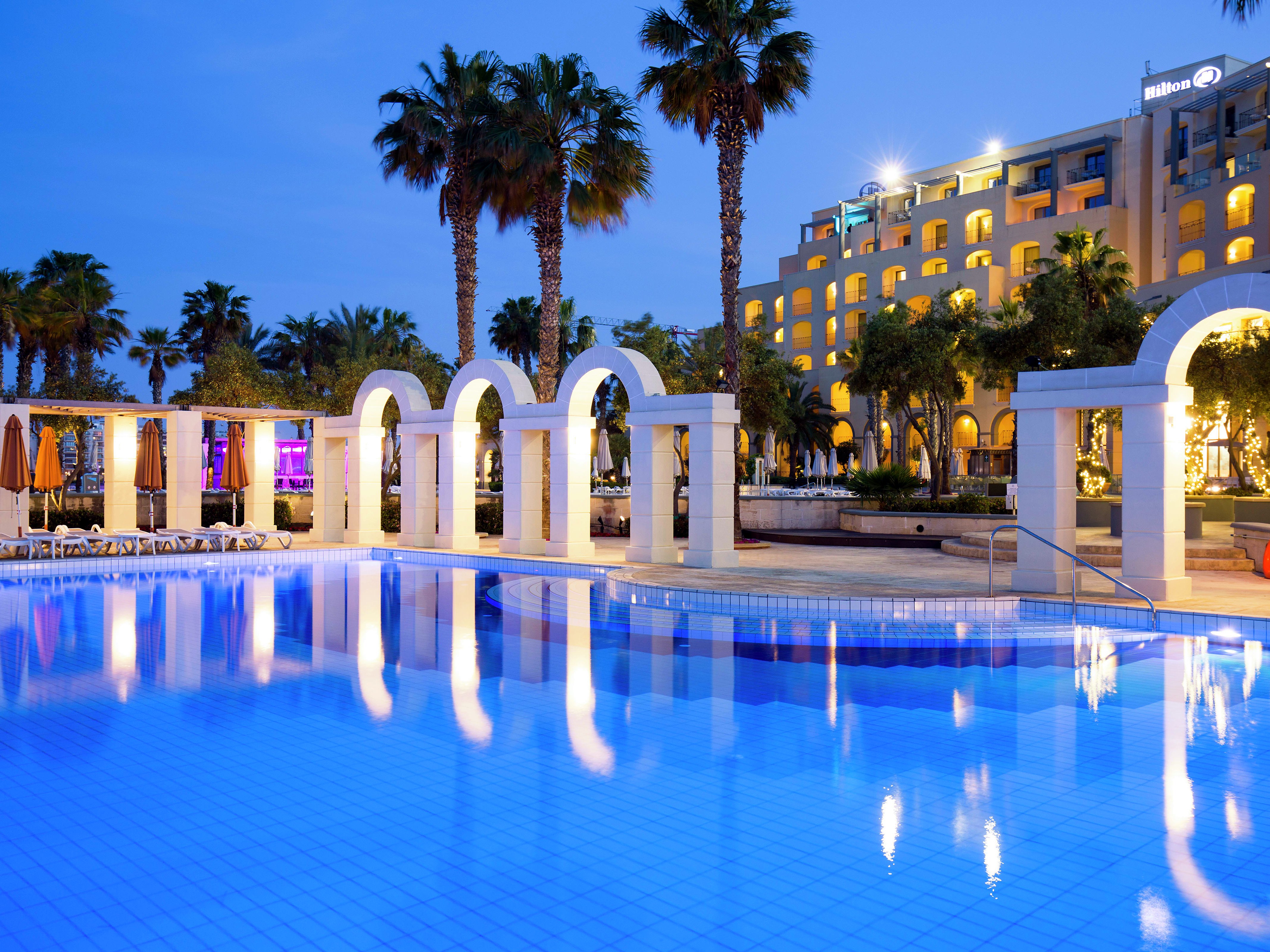 Facciata illuminata dell’Hilton Malta sopra una piscina circondata da archi e palme
