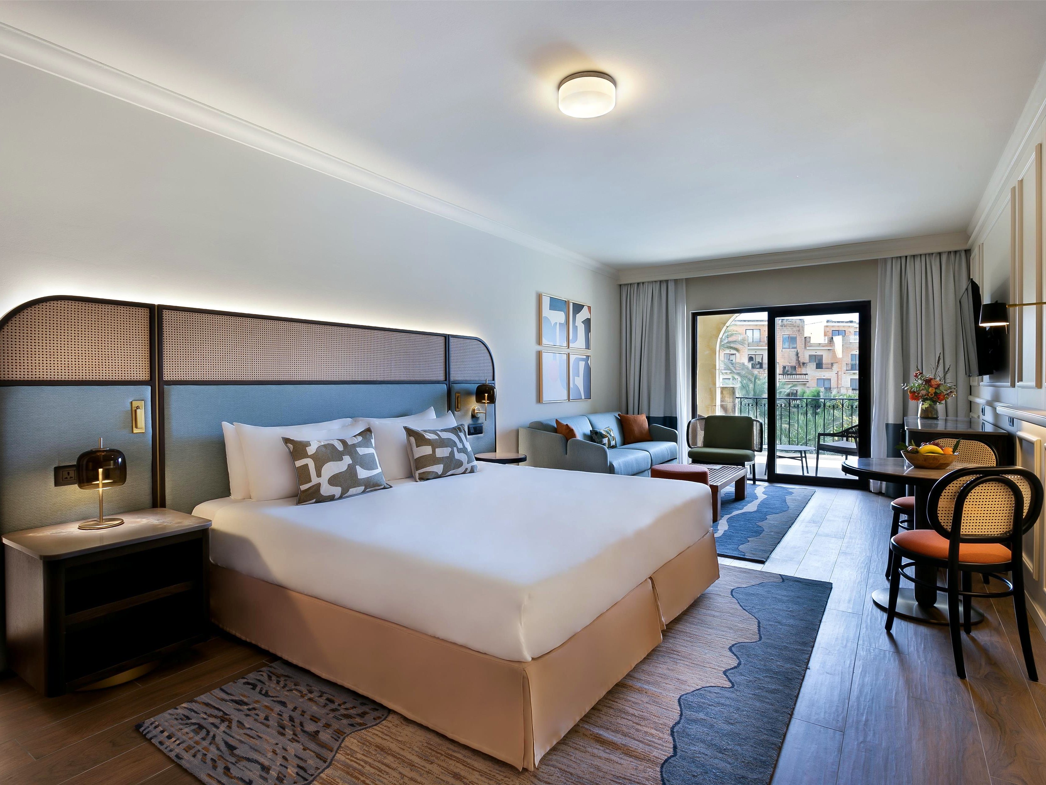 Camera Grand Deluxe vista piscina con letto matrimoniale, angolo salotto e portafinestra sul balcone al Kempinski Hotel San Lawrenz