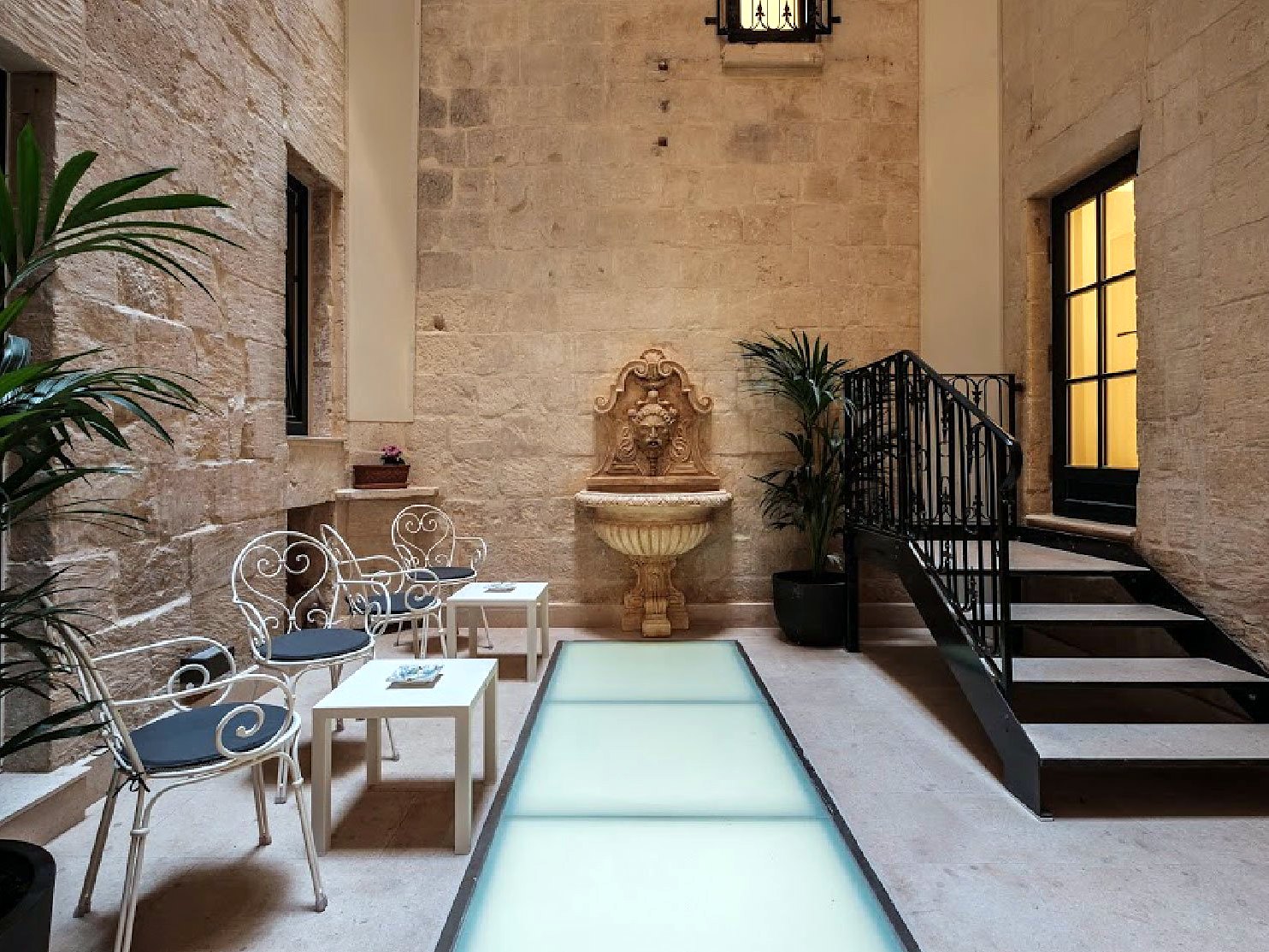 Cortile interno in pietra del Palais Le Brun con fontana a muro, scala in ferro battuto e pavimento in lastre di vetro