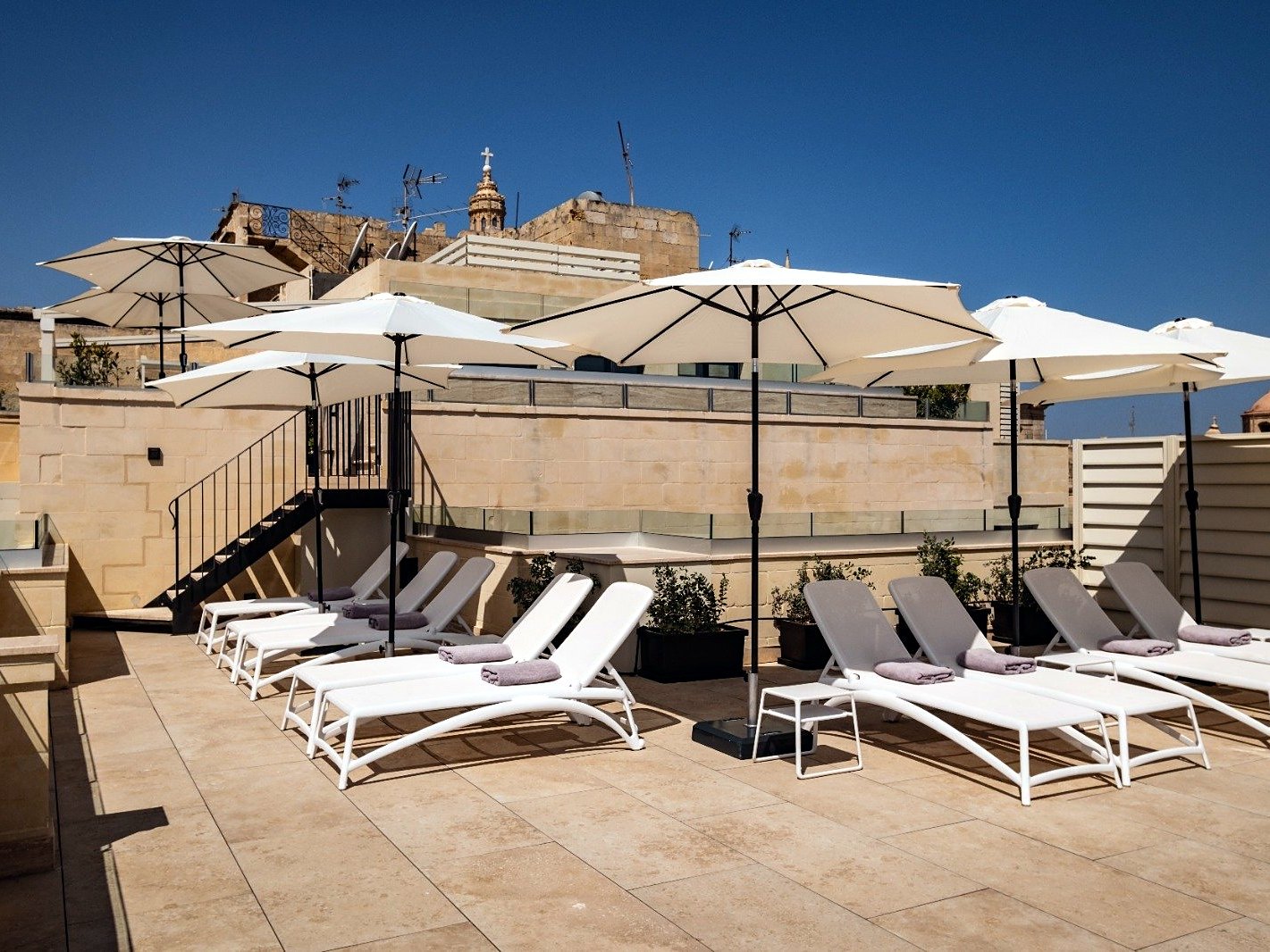 Terrazza sul tetto del Palais Le Brun con lettini bianchi e ombrelloni, sotto il campanile di una chiesa di La Valletta
