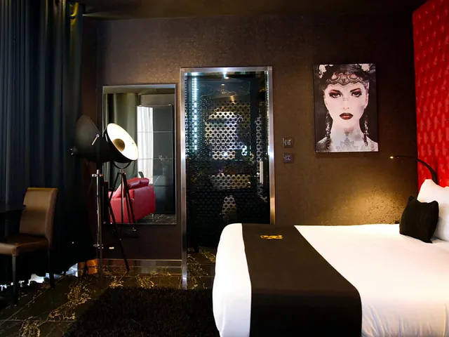 Chambre du Hugo’s Boutique Hotel avec tête de lit capitonnée rouge, portrait mural et douche vitrée