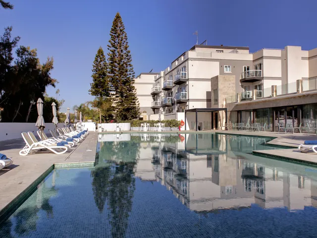 Bâtiments blancs à balcons de l’Urban Valley Resort & Spa à San Ġwann, reflétés dans la piscine extérieure bordée de transats