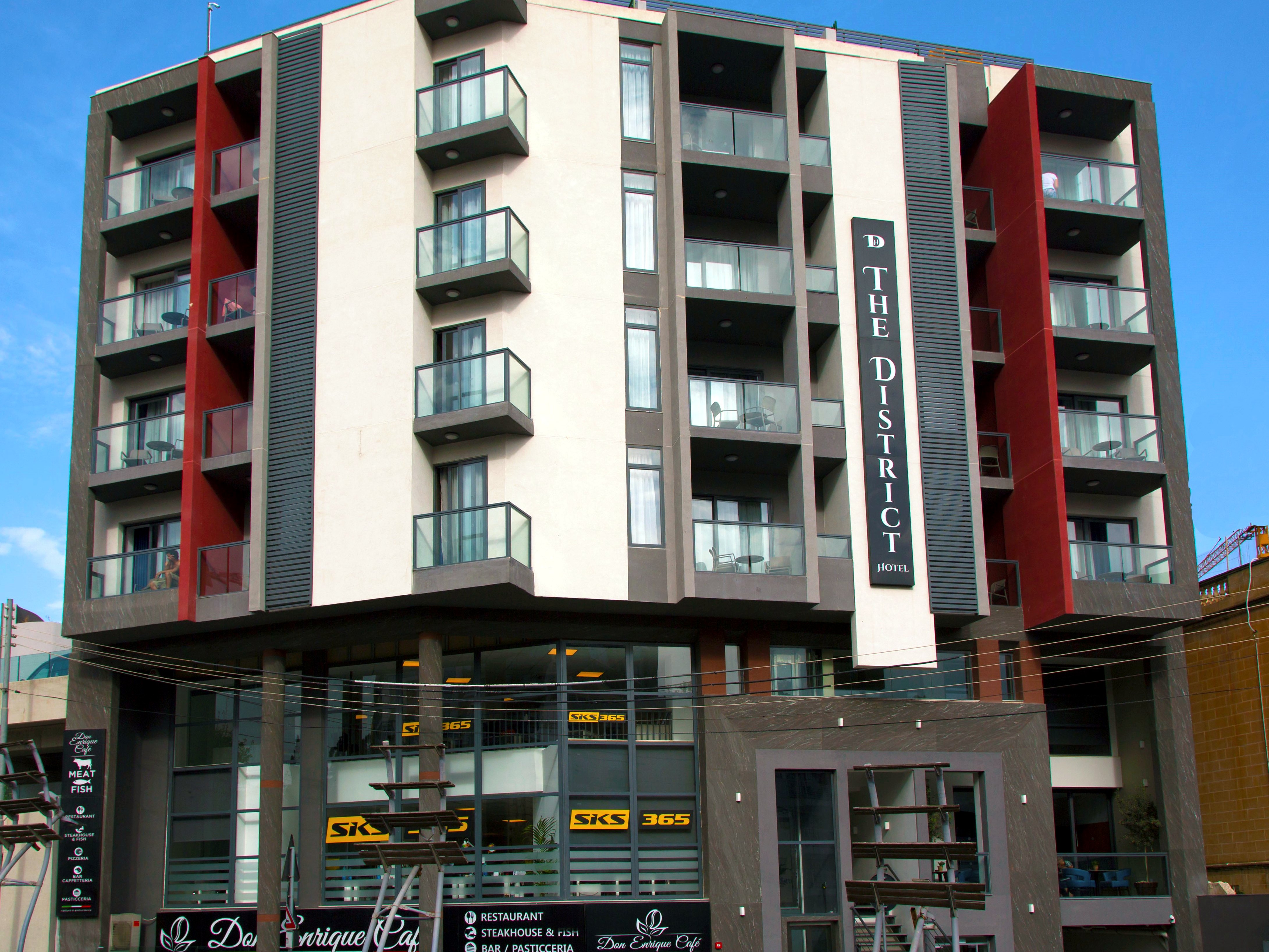 Facciata bianca e grigia di The District Hotel a St Julian’s, con balconi in vetro e insegna verticale