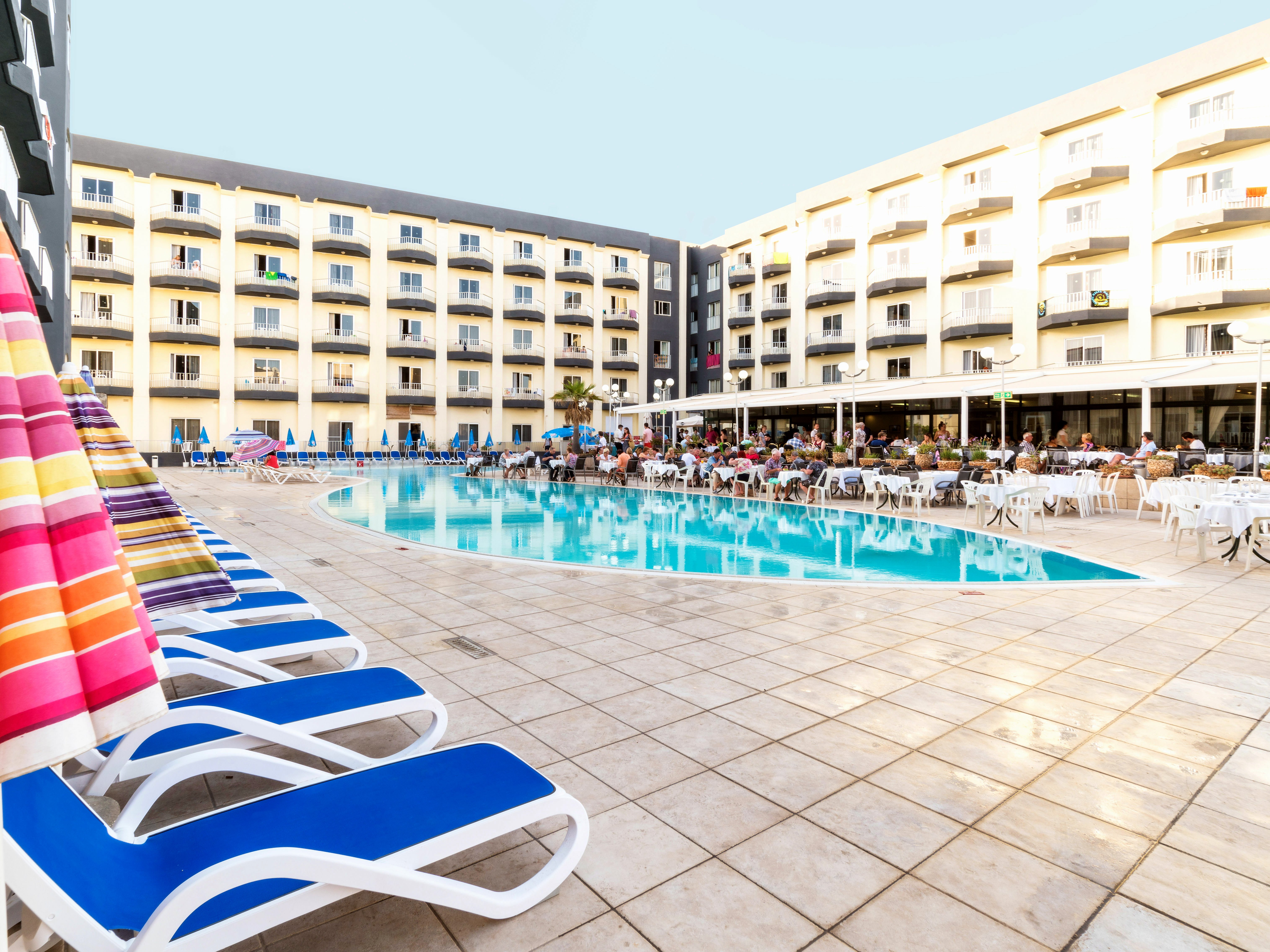 Terrazza soleggiata del Topaz Hotel con lettini blu, ombrelloni e piscina all’aperto circondata dagli edifici dell’hotel