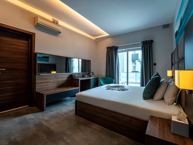 Chambre double supérieure du Pebbles Resort avec grand lit, tête de lit capitonnée, bureau et porte-fenêtre sur le balcon