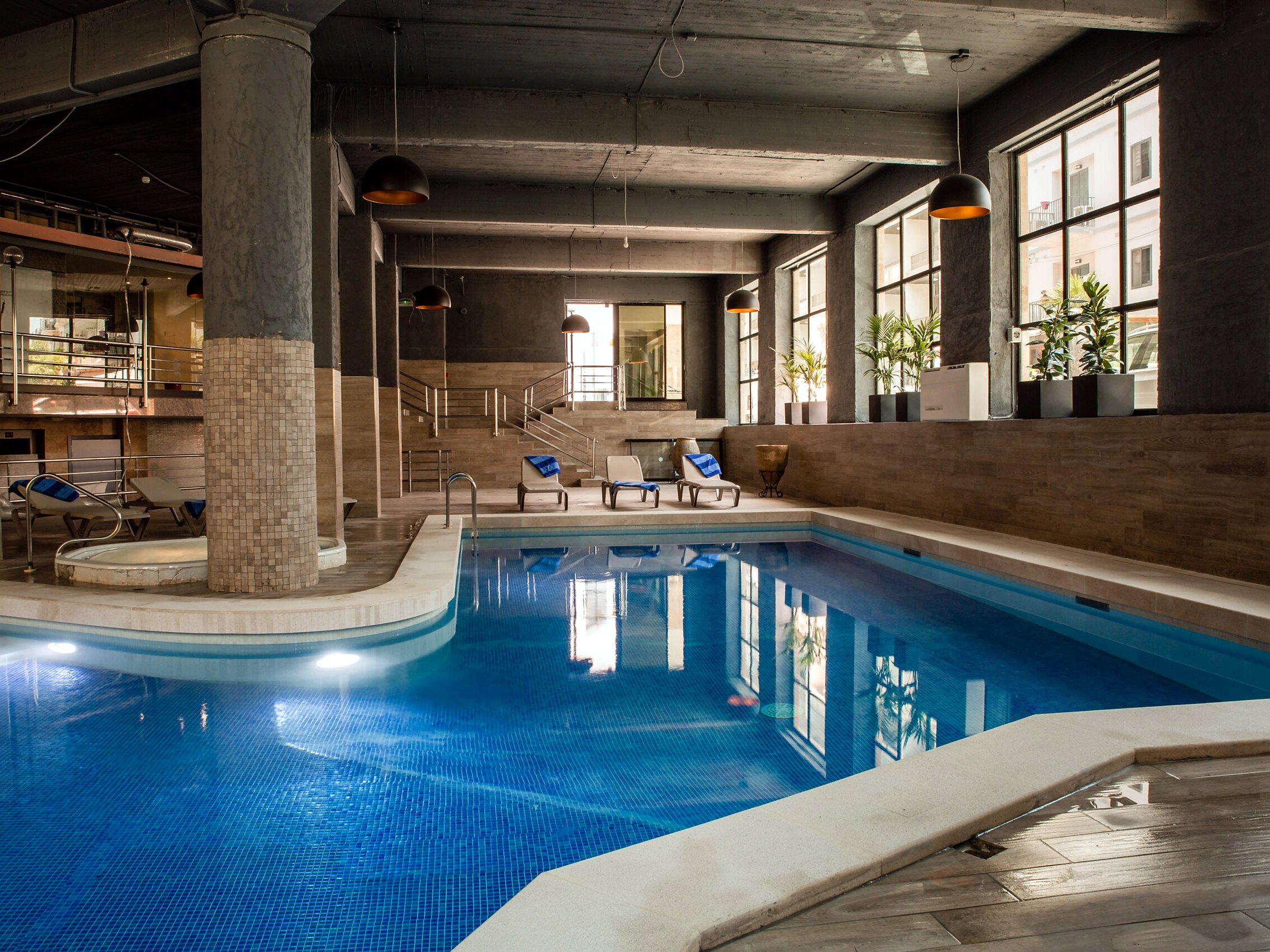 Piscina interna riscaldata del Pebbles Resort sotto un soffitto industriale, con pilastri a mosaico e lettini