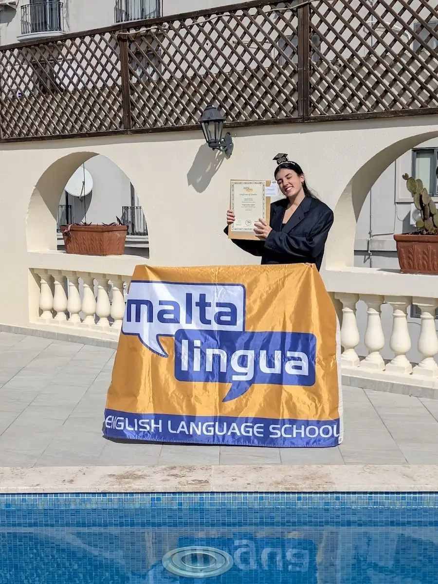 Estudiante con su diploma frente a una bandera y la piscina de Maltalingua